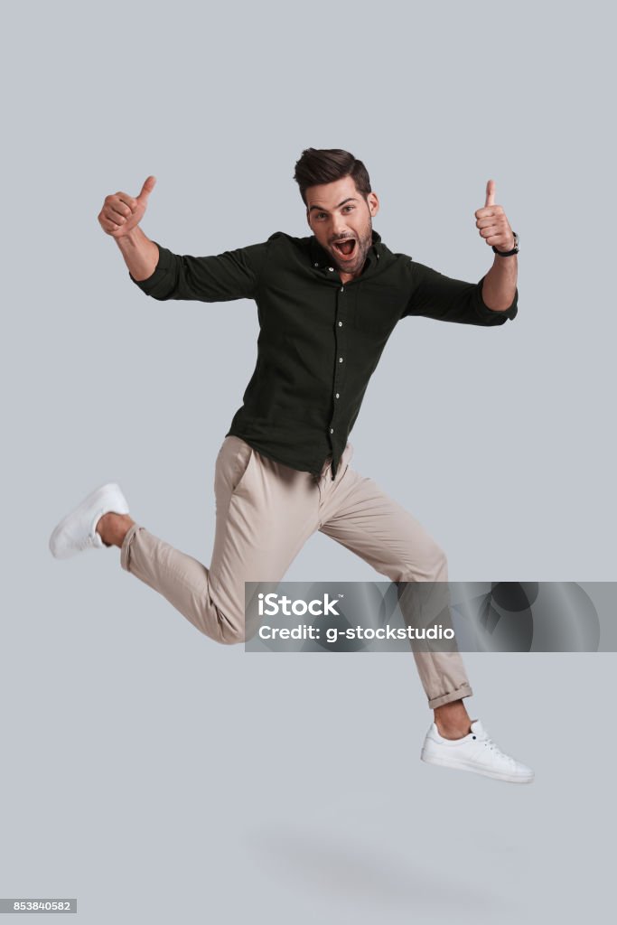 Man in mid-air. Full length of playful young man gesturing and smiling while jumping against grey background Jumping Stock Photo