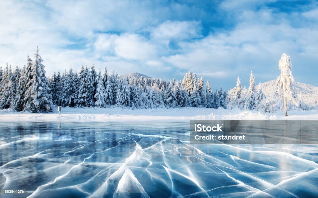 Blue ice and cracks on the surface of the ice. Frozen lake under a blue sky in the winter. The hills of pines. Winter. Carpathian, Ukraine, Europe. Blue ice and cracks on the surface of the ice. Frozen lake under a blue sky in the winter. The hills of pines. Winter. Carpathian, Ukraine, Europe Winter Stock Photo