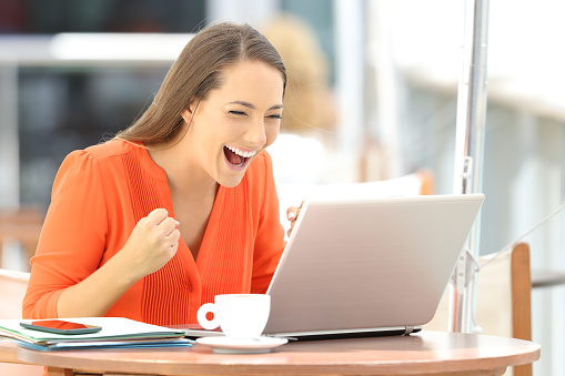 Single excited entrepreneur receiving good news on line in a laptop sitting in a restaurant terrace