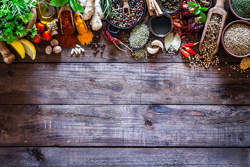 Top view of a rustic wood kitchen table with a large group of multi colored spices and herbs arranged in a row at the top border making a frame and leaving useful copy space for text and/or logo. Spices and herb included are clove, turmeric, bay leaf, cinnamon, olive oil, balsamic vinegar, curry powder, ginger, nutmeg, peppercorns, cinnamon, salt, chili pepper, basil, parsley, lemon, rosemary, garlic, onion and saffron. DSRL studio photo taken with Canon EOS 5D Mk II and Canon EF 100mm f/2.8L Macro IS USM