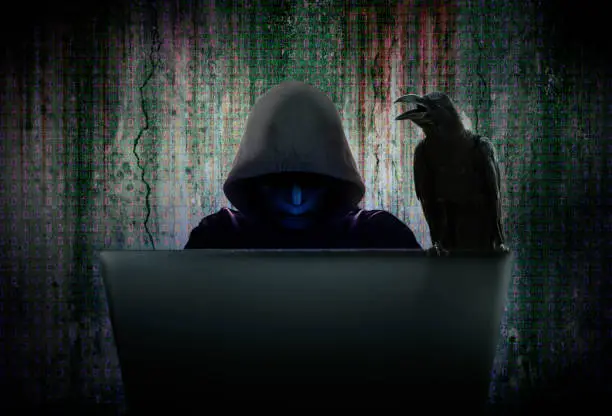 Computer cyber criminals in black mask and hood are hacking data to infiltrate and attack corporate security systems with the black crow is hanging on the computer