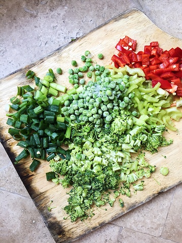 Sliced vegetables (green pepper, green onion, frozen green peas, red pepper, broccoli) laying on a cutting board ready to be cooked into healthy vegetarian dish