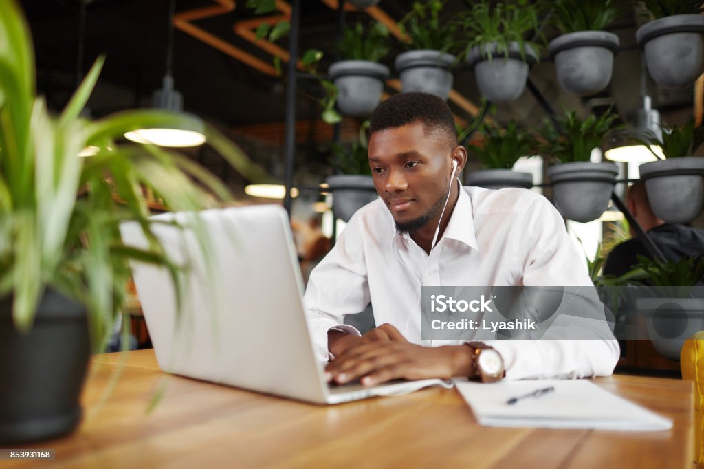 Man in headphones working at laptop in stylish cafe Serious african man in headphones, listening music, typing, working at laptop in stylish cafe. Handsome freelancer looking at screen, writing message. Student learning. Coffee break. Loft interior. Laptop Stock Photo