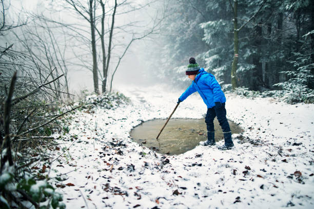 Little boy aged 7 is playing with a puddle in winter forest. The boy is breaking the ice on the puddle with stick. Simple childhood joy.\n\n
