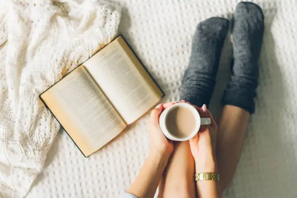 Photo of woman laying in bed and read book with cup if coffee.