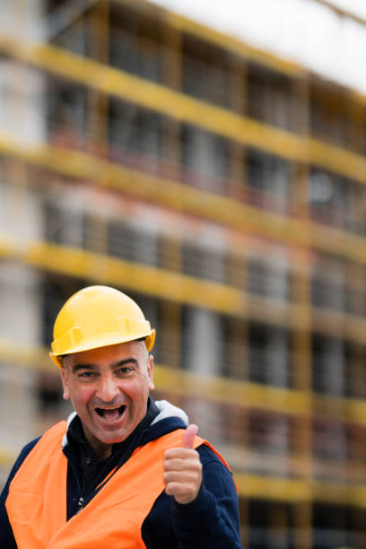 Cheerful and successful construction worker posing showing thumbs up gesture. Outdoors. Selective focus