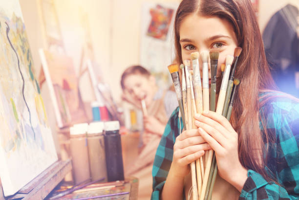 Beautiful girl holding bunch of messy painting brushes True little painter. Selective focus on a young lady of magical beauty hiding her face behind painting brushes while sitting at a painting easel and posing for the camera. painting activity photos stock pictures, royalty-free photos & images