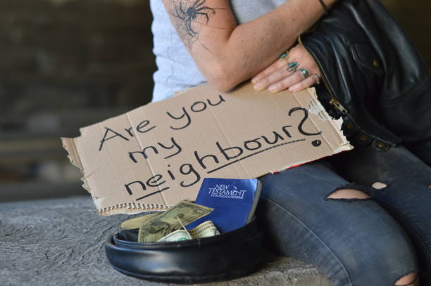 homeless tattooed person begging for money holding a sign - are you my neighbor? - with a bible in her collecting cap