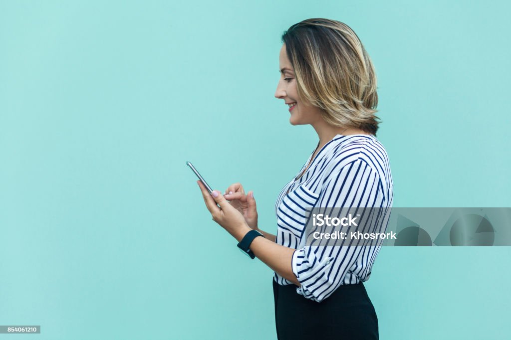 Side view of happiness business woman, punished alone at blue wall and using smartphone. Side view of happiness business woman, punished alone at blue wall and using smartphone. Indoor studio shoot Colored Background Stock Photo