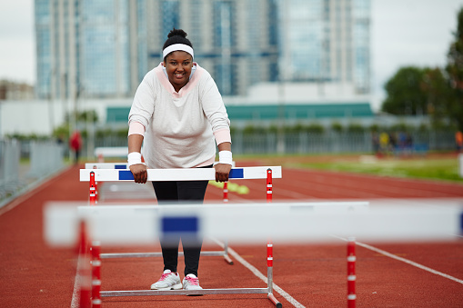 Happy plus-sized woman in activewear leaning forward on hurdle during training on stadium