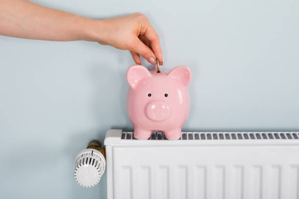 Woman Hand Inserting Coin In Piggybank On Radiator Close-up Of A Woman Hand Inserting Coin In Piggybank On Radiator At Home radiator heater stock pictures, royalty-free photos & images