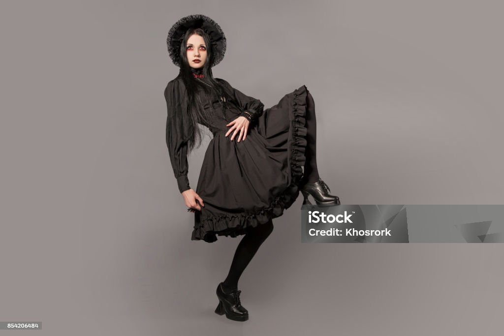 Portrait of brunette woman in black dress and classic gothic style with red eyes on gray background. halloween concept. Goth Stock Photo