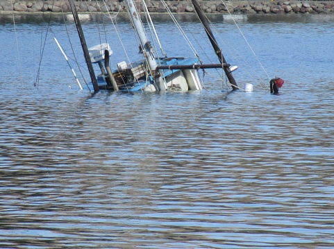 A Boat is sinking almost completely underwater not far from a rocky shoreline on a sunny day.