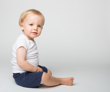 A 15 moth old blonde haired boy photographed while sitting in a white photo studio while wearing a white t shirt and blue shorts.