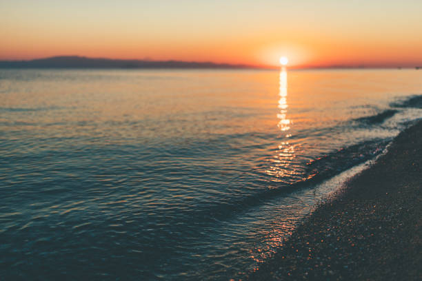 Long exposure photo of a wind-swept lake during sunset, where the motion of the waves creates a smooth, misty surface. In the background, gentle hills stretch across the horizon under a softly glowing sky. The image conveys a peaceful, dreamy atmosphere, ideal for concepts such as serenity, nature's beauty, calmness, and scenic landscapes.
