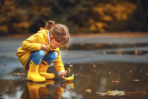 Photo of happy child girl with umbrella and paper boat in   puddle in   autumn on nature