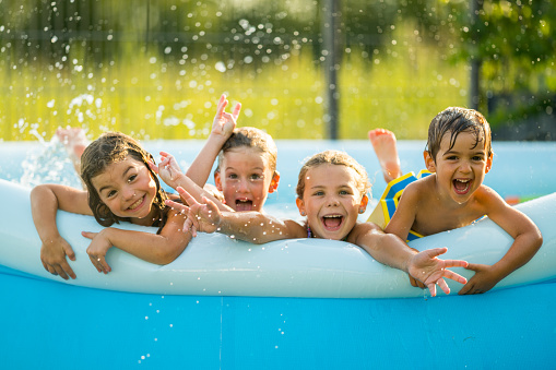 four young children kids siblings at the age of 3, 4 and 6 in blue inflatable swimming wading paddling pool with wet hair laughing smiling enjoying the cooling in heat wave shallow focus water drops giving the feeling of refreshing