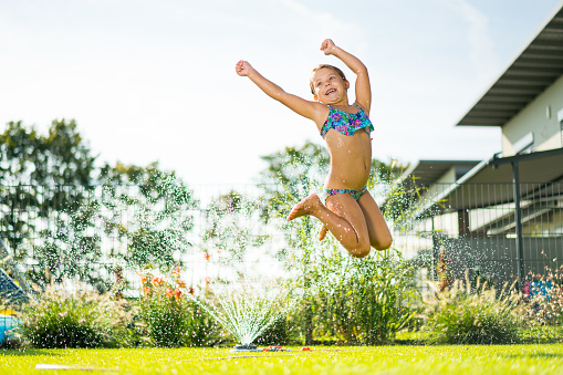 6 year old girl playing with and jumping over garden sprinkler in grass backyard of modern house backlight making the water drops visible