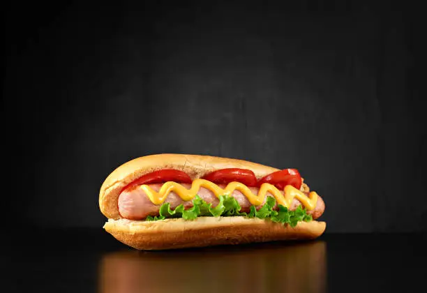 Big hotdog with sausage tomatoes, mustard and salad isolated on black background. Top view.