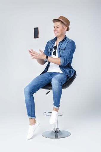 handsome man in denim sits on chair with phone on white background