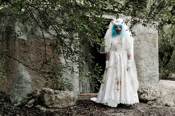 scary evil clown in a bride dress a scary evil clown wearing a dirty and ragged bride dress, at the door of a rustic shelter, in a disturbing rural landscape scary bride stock pictures, royalty-free photos & images