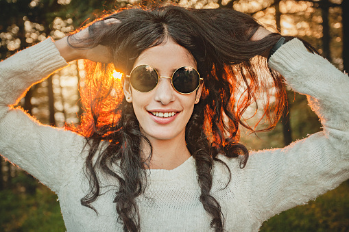 Outdoor image of cool, happy beautiful Asian young woman holding her hair and enjoying in nature at the time of sunset. She is wearing circular sunglasses and looking at camera with toothy smile. One person, waist up and selective focus. Image shot in Sirmaur, Himachal Pradesh.