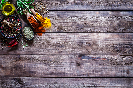 Top view of a rustic wood kitchen table with some spices and herbs arranged at the top-left corner leaving useful copy space for text and/or logo. Spices and herb included are turmeric, olive oil, curry powder, peppercorns, chili pepper, basil and rosemary. DSRL studio photo taken with Canon EOS 5D Mk II and Canon EF 100mm f/2.8L Macro IS USM