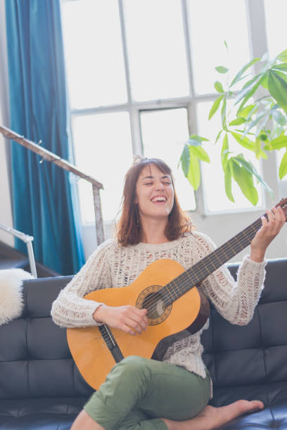 istock Beautiful young woman sitting on sofa playing guitar 854405414