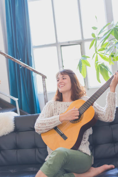 istock Beautiful young woman sitting on sofa playing guitar 854408580