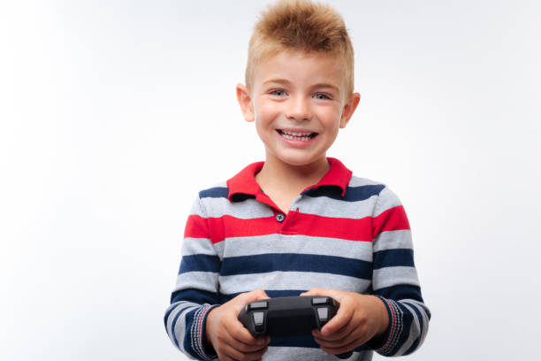 Favorite toy. Upbeat fair-haired little boy holding a game controller while playing a video game and smiling happily