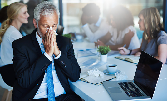 Shot of a frustrated businessman using a tissue to sneeze in while being seated in the office