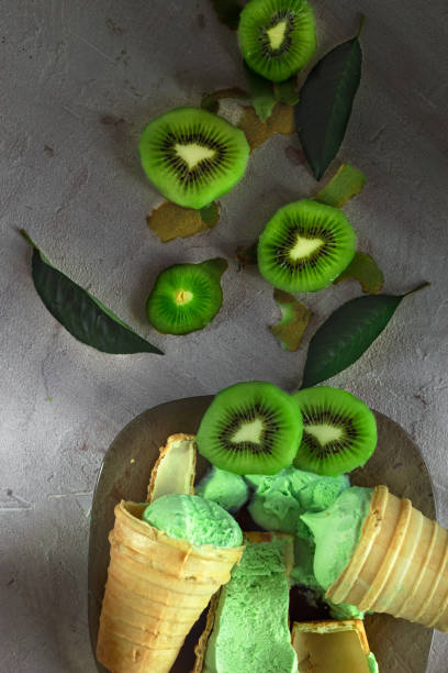 istock Ice cream green in a cone, three pieces, one is cut. Kiwi is cut with segments, on a black plate. Vertical. Top view. 854441838