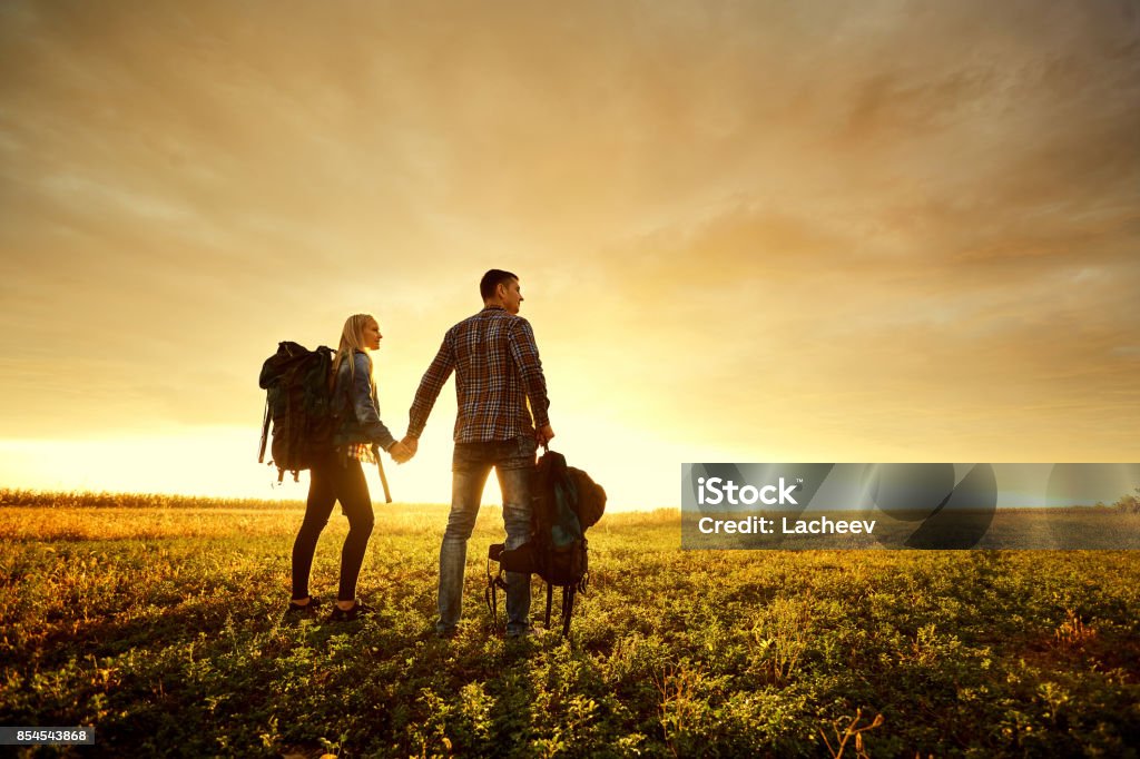 Um casal de turistas com mochilas na natureza ao pôr do sol. - Foto de stock de Adulto royalty-free