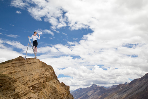 Businesswoman standing on ladder and shouting through a megaphone on top of cliff