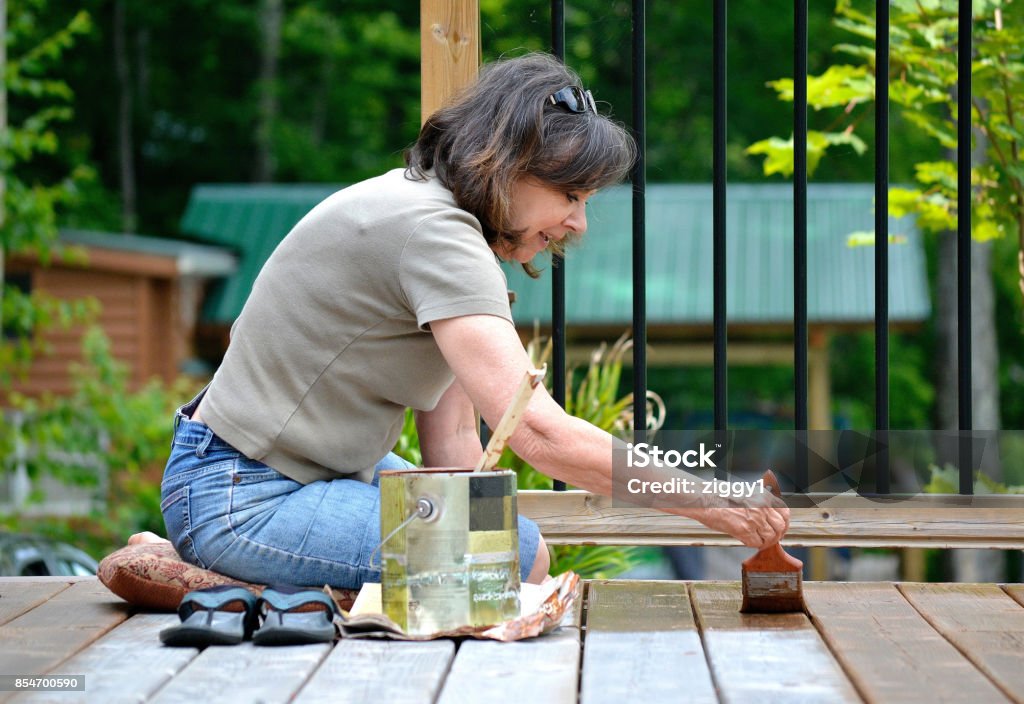 Lifestyle, Senior woman, 50+, paints a deck. This an image of a female senior, 50+, staining by hand, with a brush, a large deck at a summer cottage. Deck Stock Photo