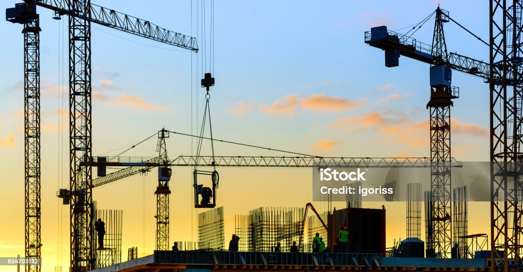 Grúas de torre y edificio de silueta con trabajadores al atardecer. - Foto de stock de Solar de construcción libre de derechos