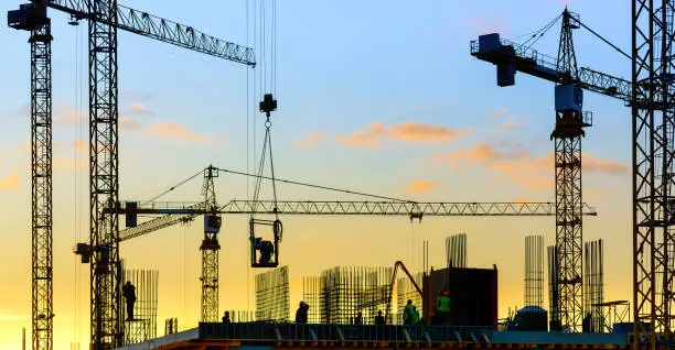 Photo of Tower cranes and building silhouette with workers at sunset.