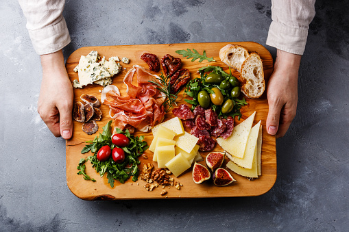 Italian snacks food with Ham, Olive, Cheese, Sun-dried tomatoes, Sausage and Bread on wooden cutting board in male hands on concrete background