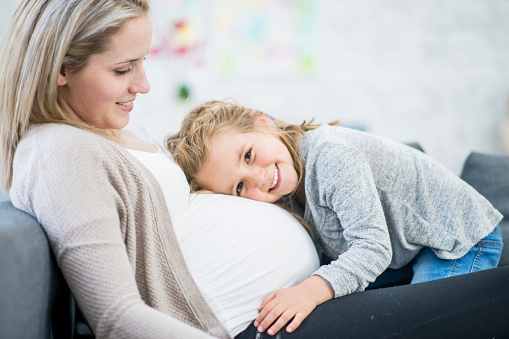 A pregnant Caucasian mother and her daughter are indoors in a living room. They are wearing casual clothing. The daughter is laying her head on her mother's stomach.