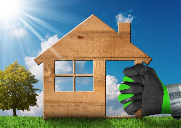 Hand with work glove holding a wooden model house on green grass with a tree and a blue sky with clouds and sun rays. Concept of ecological house