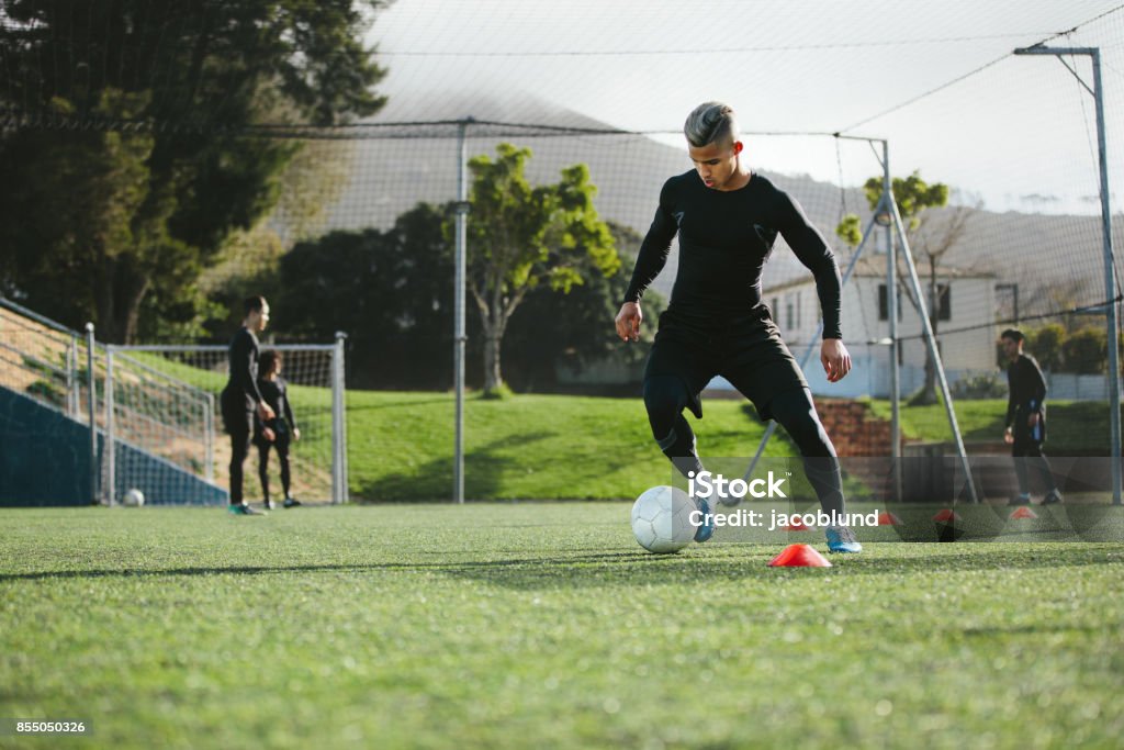 Football player practice dribbling on field Young soccer player training in football field with team in background. Five a side football team practicing on field outdoors. Soccer Stock Photo