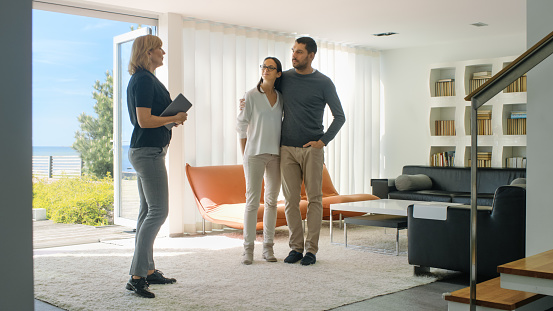 Professional Real Estate Agent Shows Stylish Modern House to a Beautiful Young Couple Who are in the Market for Purchasing/ Renting New Home. House Has Floor to Ceiling Windows and Seaside View.