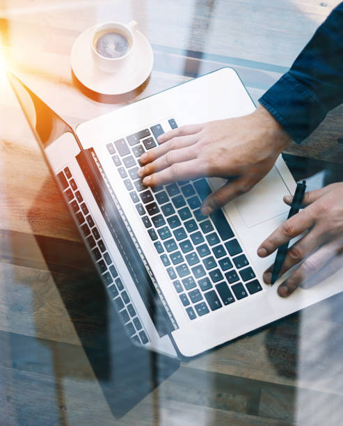 Man working on laptop at sunny office.Male hand typing on keyboard.Modern notebook, cup of black coffee on the wooden table.Reflections on glass surface.Vertical.