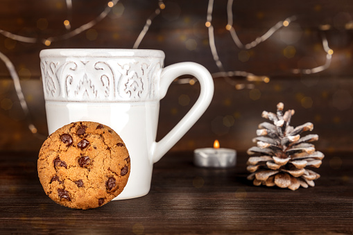 A photo of a chocolate chips cookie with a cup of hot chocolate behind it, with a snowy pine cone, a candle, and fairy lights in the blurred background, with copy space, side view, on dark textures