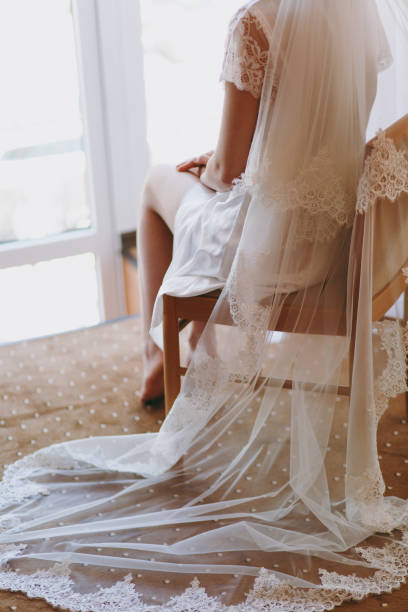 istock Bride brunette in veil and white satin dressing gown sits by the window 855172938