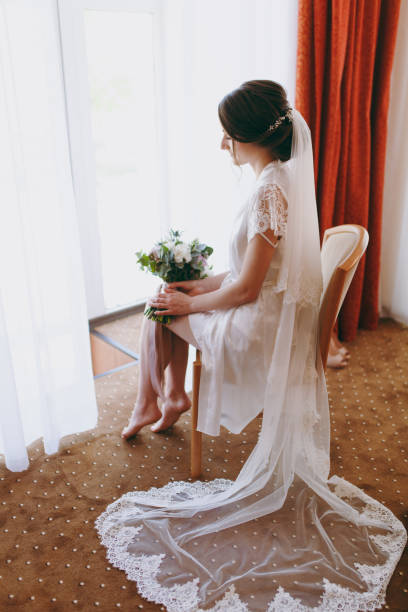 istock Bride brunette in veil and white satin dressing gown sits by the window 855175970