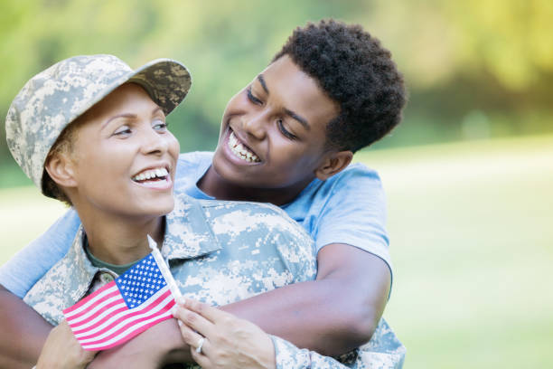 Teenage son and military mom are reunited after her tour of duty A handsome African American teenage boy stands behind his military mom, dressed in camo, and smiles at her as he wraps his arms around her. She is holding a small american flag as she turns to smile back at him. proud army mom stock pictures, royalty-free photos & images