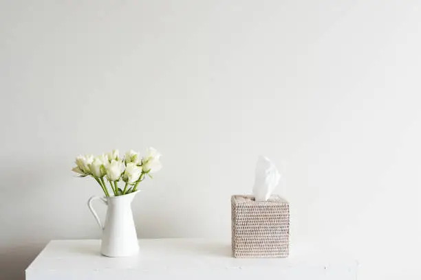White roses in jug on shelf with rattan tissue box against neutral wall background