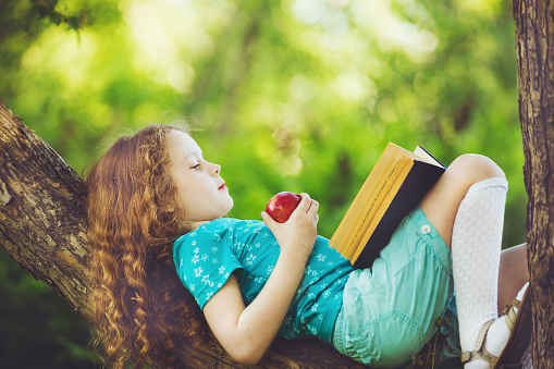 Little girl lying on large tree, eats red apple and reads the book.