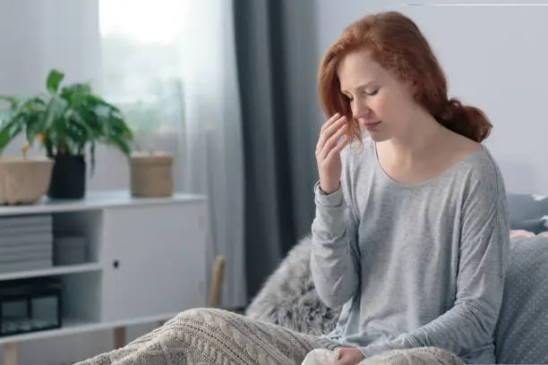 Young woman sick with flu having headache sitting on the bed suffering from runny nose and high fever